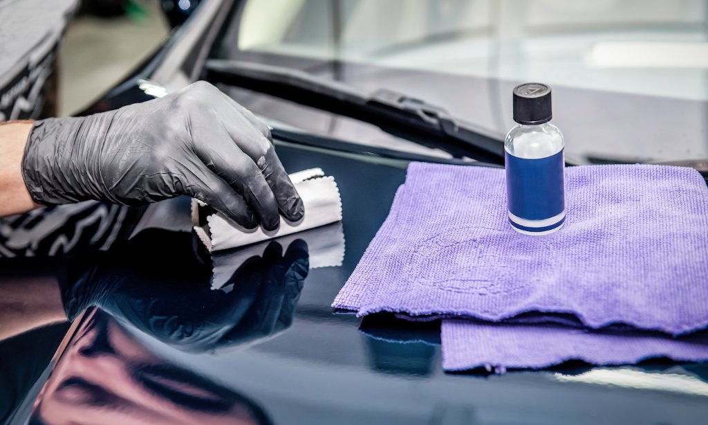 Gloved hand applying paint protection to a car hood next to a bottle and folded microfiber towels